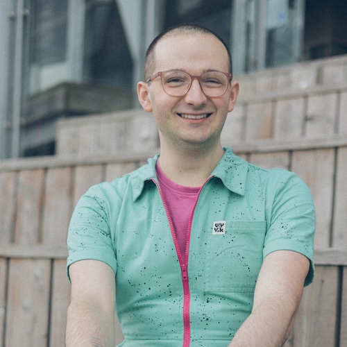 Andrew Houghton, a 27 year old non-binary individual wearing a turquoise jumpsuit with a pink T-shirt and glasses. They are smiling directly into the camera.