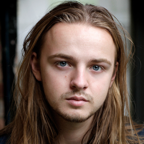 Charlie, a white man, with shoulder length dark blonde hair in a windswept style. He has blue eyes and is wearing a navy t-shirt. He has a stubble. He is looking at the camera with a poised expression.