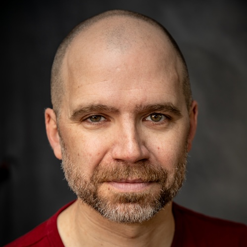 Dan McGarry, a white male in his early 40s with a shaved head and closely cropped greying beard looks at the camera with his brown eyes and a half-smile. He wears a maroon t-shirt.