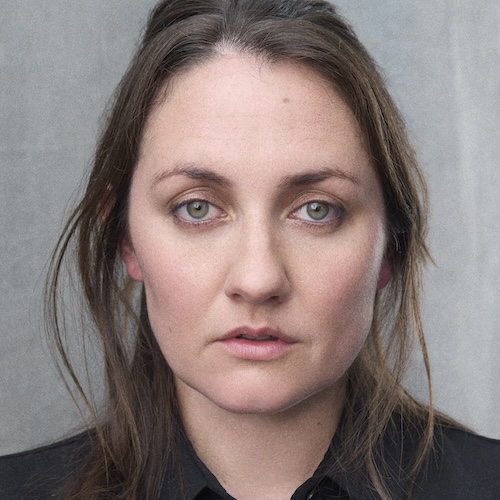 Emma Bright, a white woman, with long brown hair half up half down, wearing a black shirt stares straight in to the camera with a neutral expression. She has green eyes.