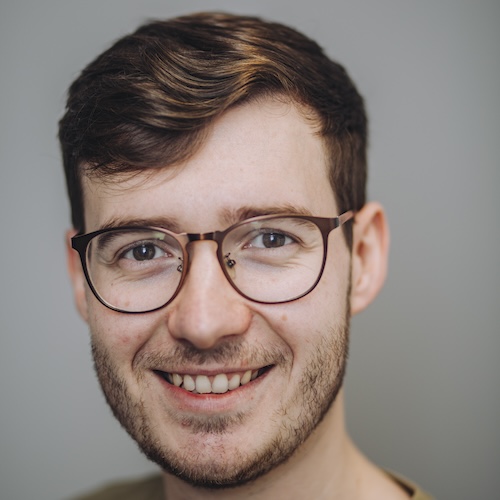 Jack Chamberlain, a white man of slim build with brown hair wearing glasses and a green t-shirt smiles at the camera. He has hazel eyes.