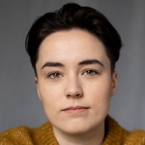 Jo, a white, androgynous looking, non-binary woman with short dark brown hair and hazel eyes, looks into the camera with a gentle closed-mouth smile. She is wearing a mustard coloured knitted jumper against a dark grey background.