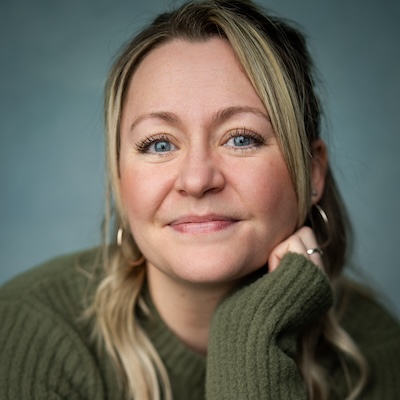 Josie Morley, a white woman of slim build with blonde hair and blue eyes, is smiling at the camera. She is wearing a green jumper and is resting her head on her hand.