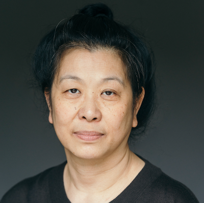 A British East and Southeast Asian woman, of medium build, black hair with a few visible grey strands, long hair done up in a bun, wearing a black round neck top. She has dark brown eyes and is smiling slightly.