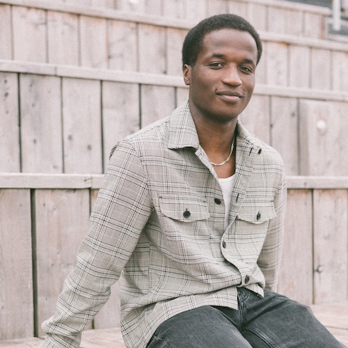 Prince Kundai, a Black man in his twenties with short black hair, a grey plaid shirt with white vest underneath and black washed denim jeans.