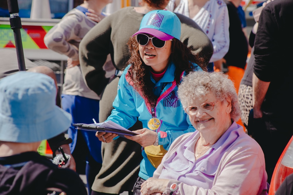 A volunteer in a blue jacket and blue baseball cap holds a clipboard while speaking with two older people.